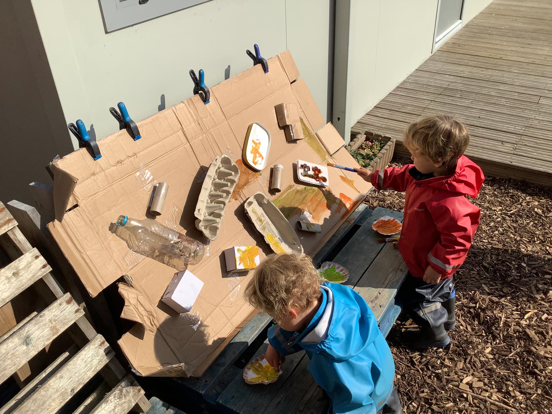 Nursery children in red and blue jackets painting cardboard boxes, tubes, and plastic bottles clamped to a wooden easel.