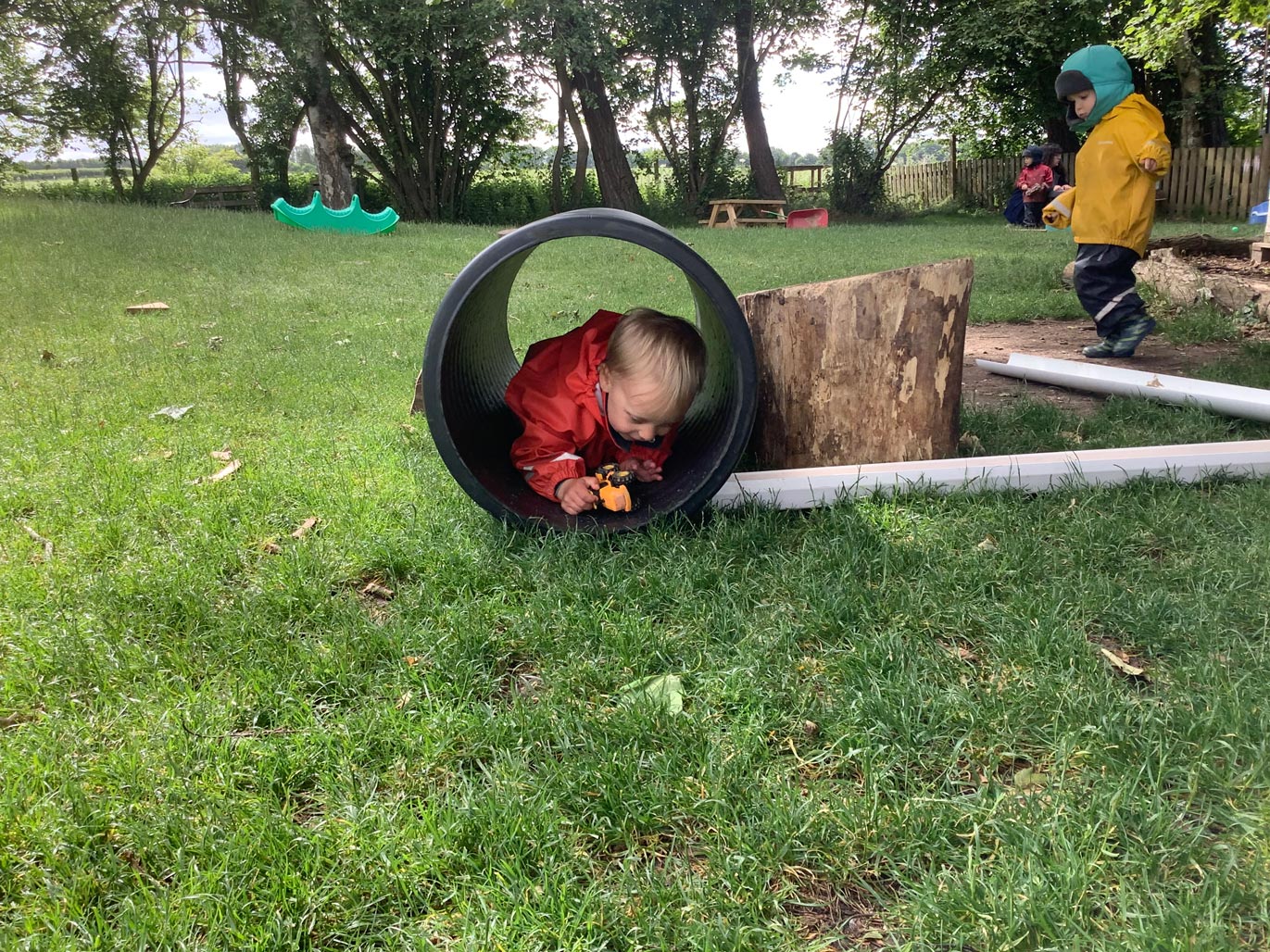 Nursery child crawling inside a black drainage pipe tunnel on a grassy outdoor play area.