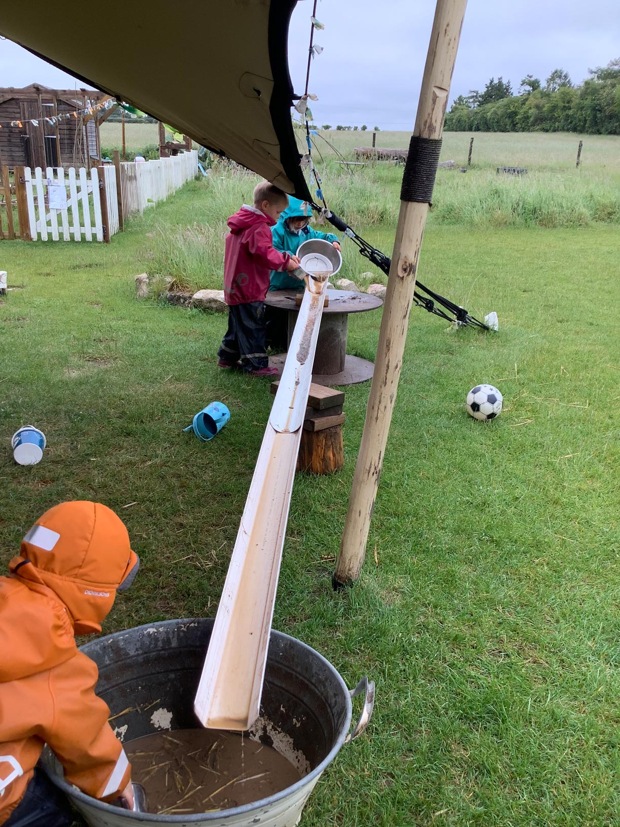 Nursery toddlers in rain jackets pouring water down a gutter channel into a tub on a grassy lawn.