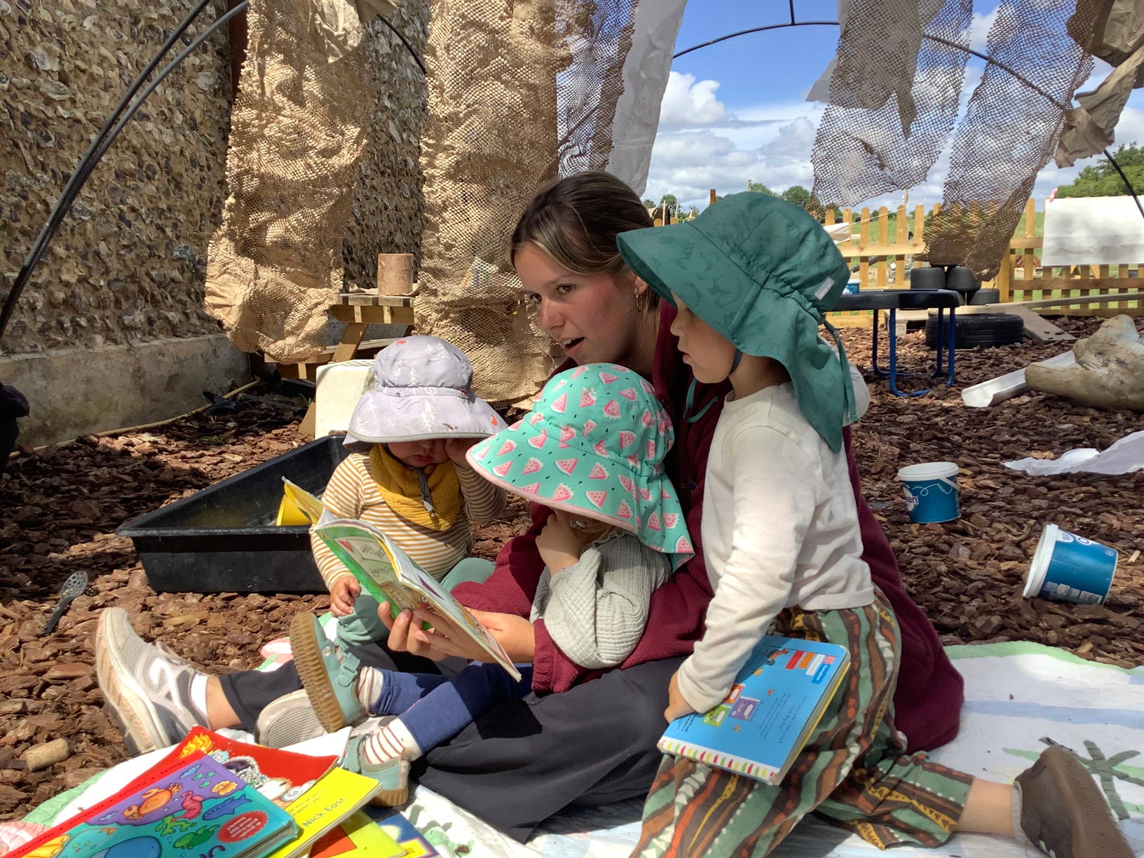 Early years practitioner reading a picture book to toddlers under a shaded outdoor canopy.