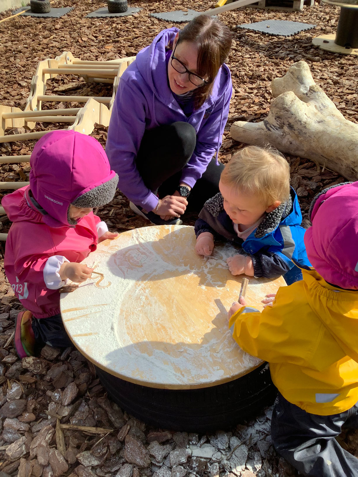 Young nursery children making marks in flour with sticks on a round table set up in a bark mulch play area.