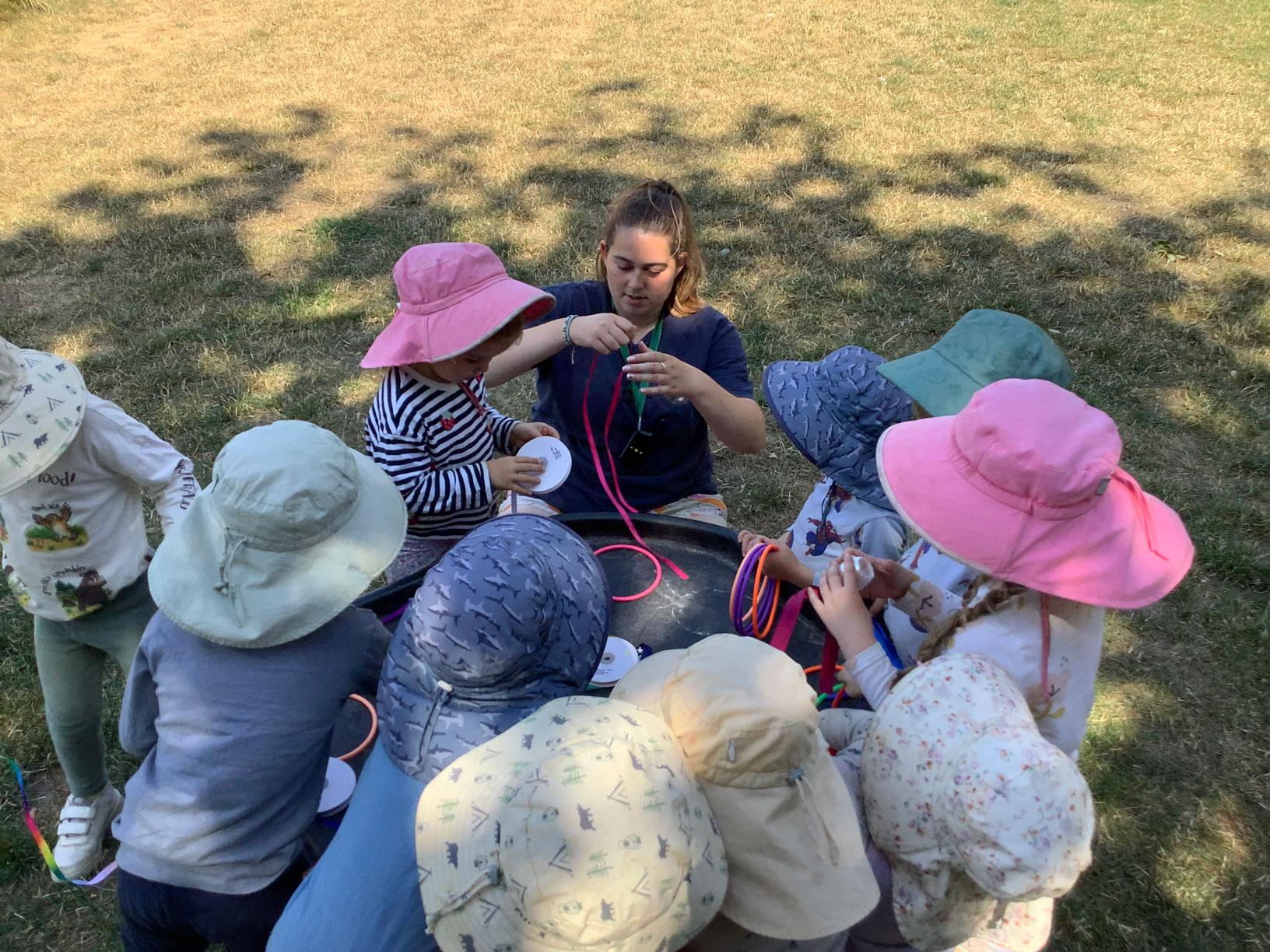Early years practitioner helping toddlers tie colourful ribbons and hoop rings around a tuff tray outdoors.