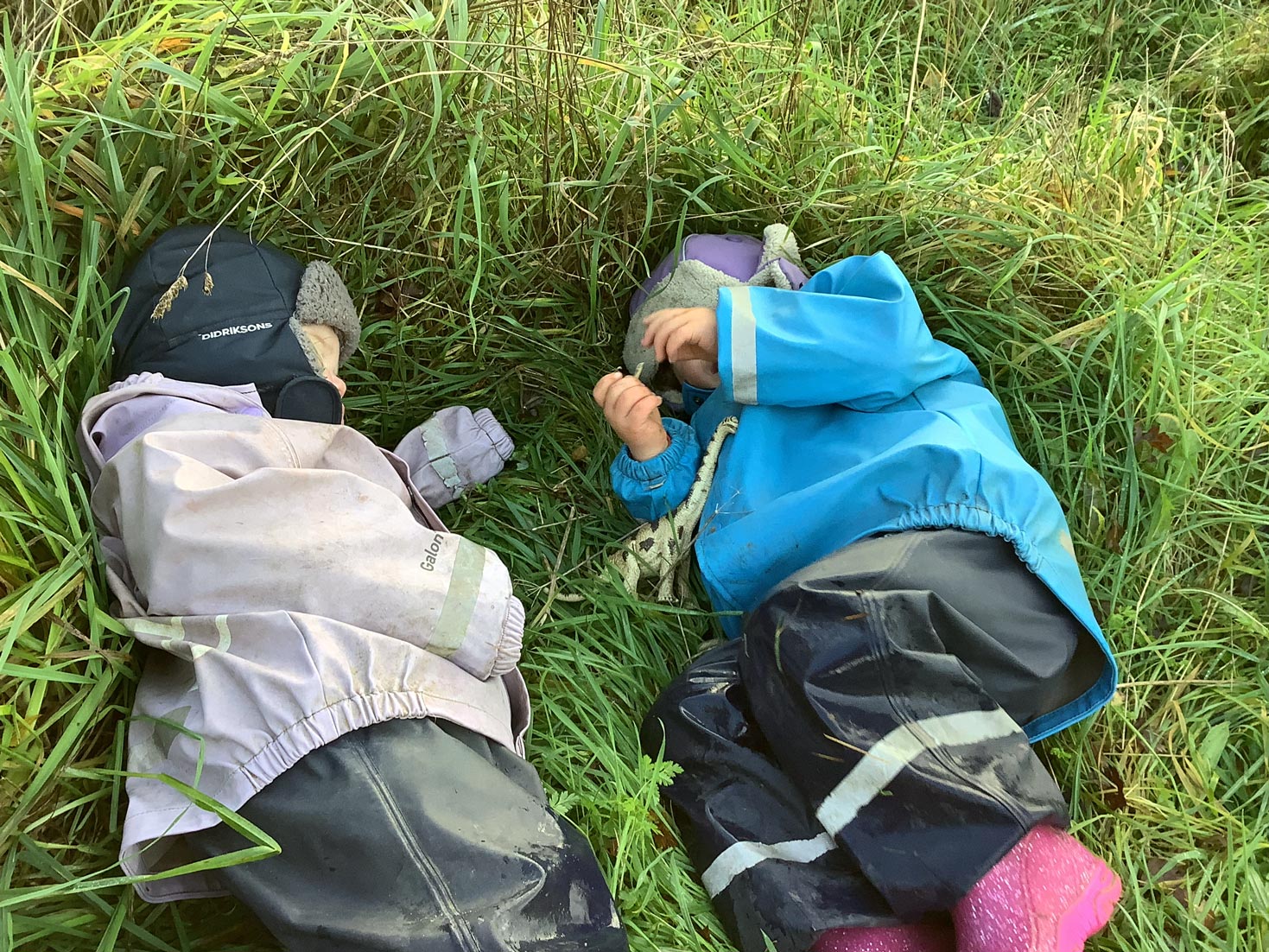 Two nursery children in rain suits resting comfortably together in tall grass during forest school play.