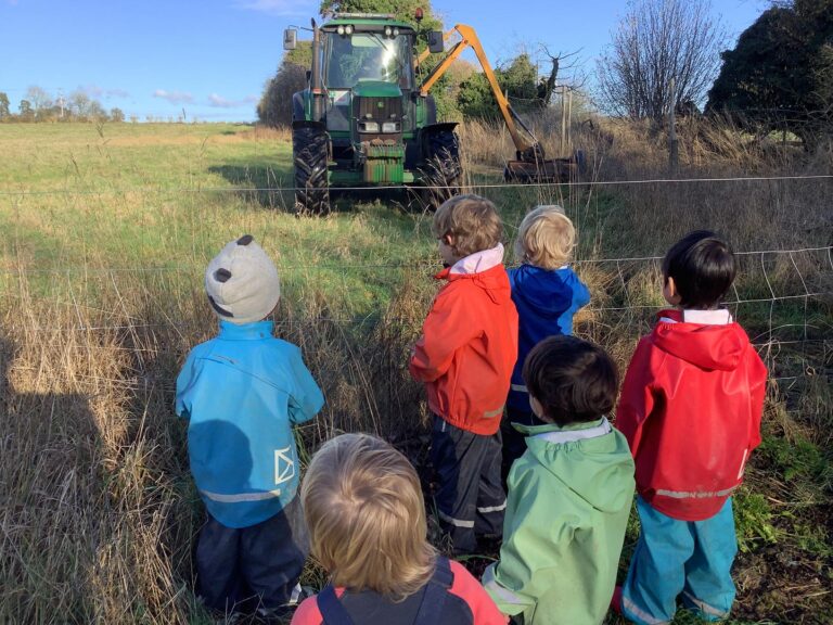 Group of nursery toddlers in bright waterproof jackets watching a John Deere tractor operate outdoors on a sunny day.