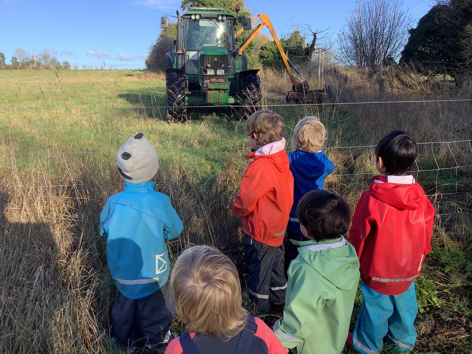 Group of nursery toddlers in bright waterproof jackets watching a John Deere tractor operate outdoors on a sunny day.