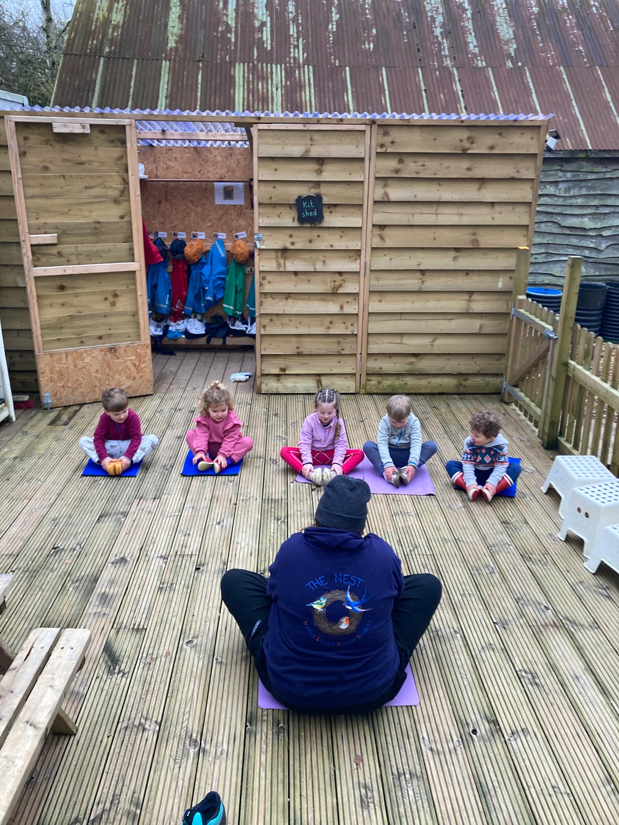 Group of nursery children following an adult instructor doing seated stretches in a circle on an outdoor decking area.