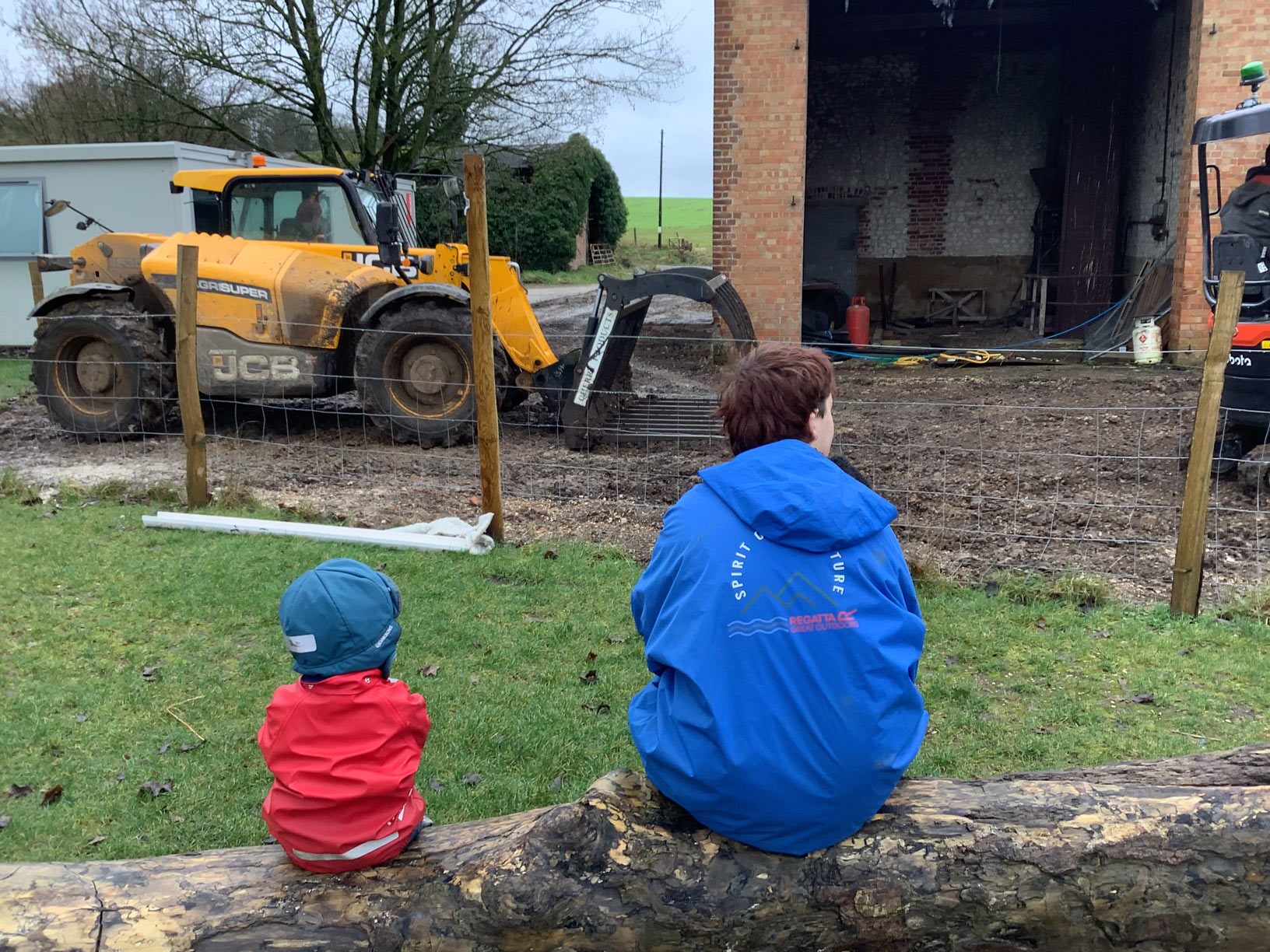 Child and adult sitting together on a log perch to watch farm and construction vehicles operating in a muddy yard.