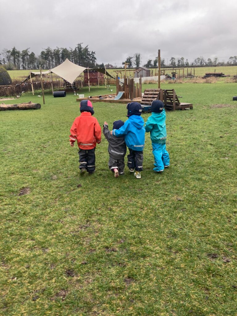 Four nursery toddlers dressed in winter rainwear walking arm-in-arm toward an outdoor play area on an overcast day.