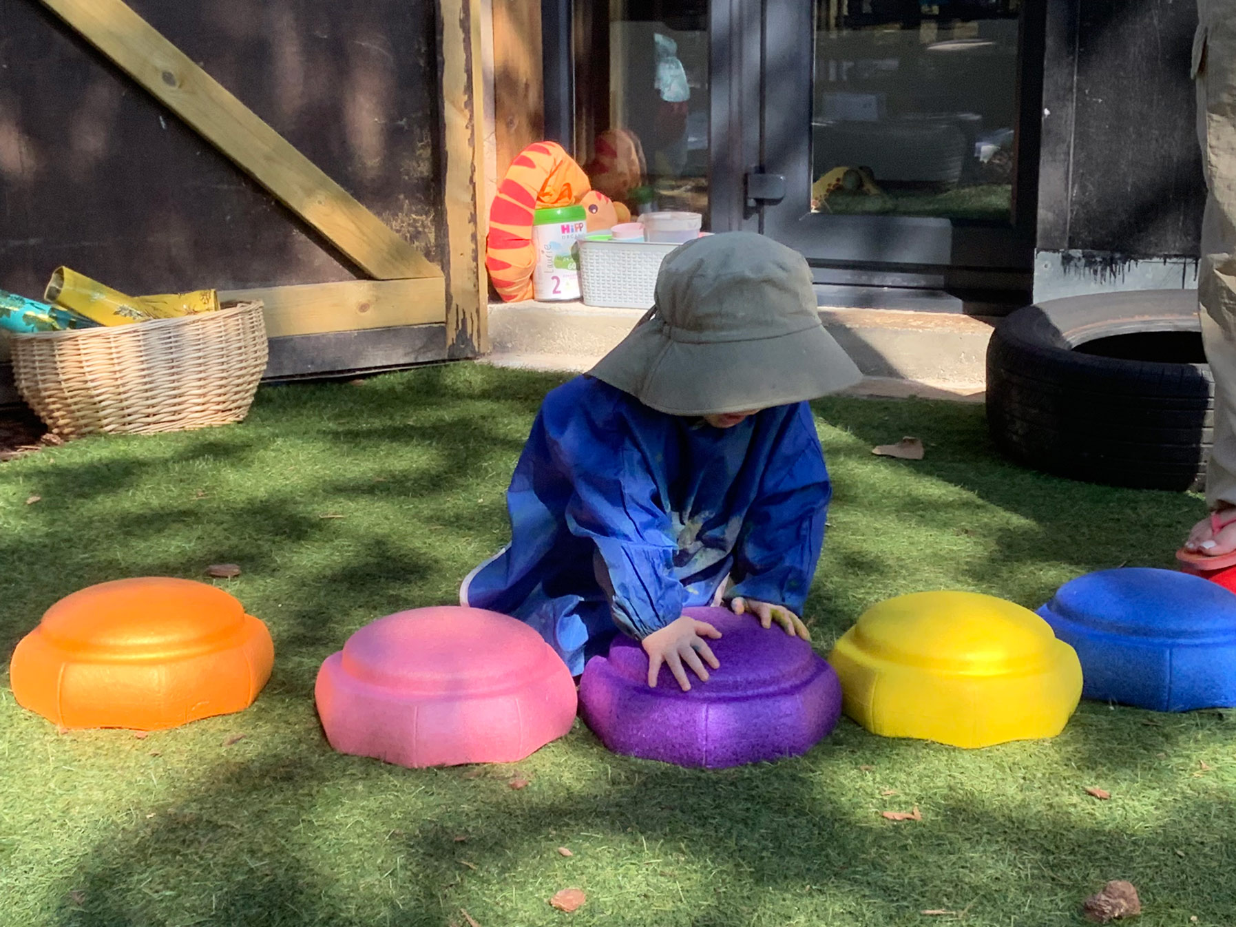 Early years toddler in a wide-brim hat and smock kneeling on artificial grass to press colourful stepping stones.