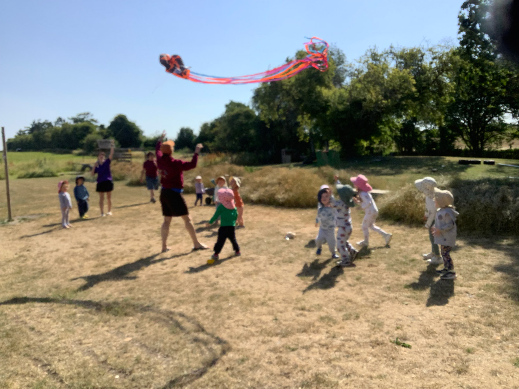 Group of nursery children in sun hats watching a large streamer kite catch the wind in a field.