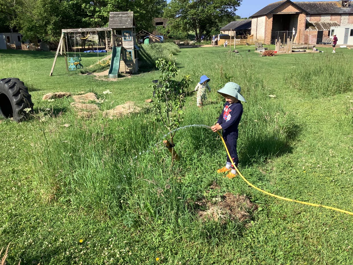 Nursery children in sun hats perched on a wooden log during outdoor play in a grassy meadow.