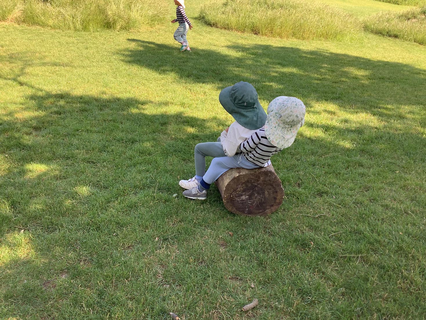 Nursery children in sun hats perched on a wooden log during outdoor play in a grassy meadow.