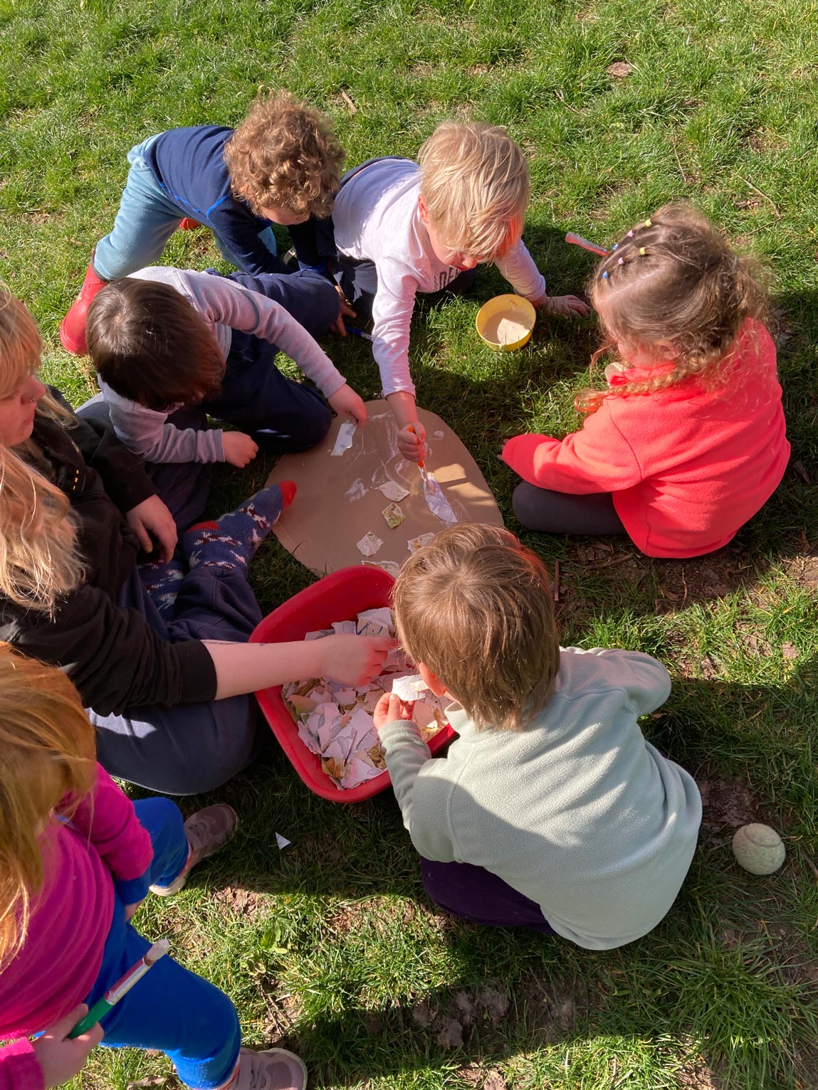 Nursery children exploring natural materials, dough, and wooden discs set up on an outdoor tuff tray on a sunny lawn.