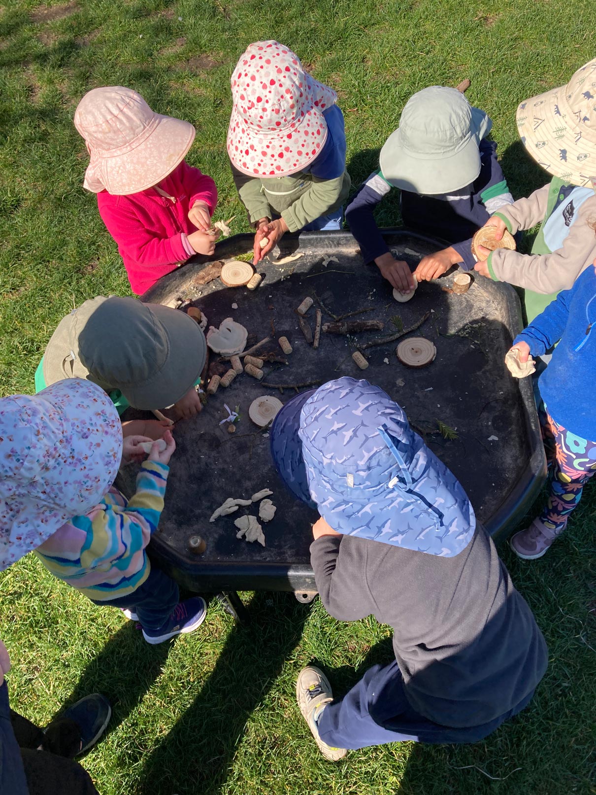 Nursery children exploring natural materials, dough, and wooden discs set up on an outdoor tuff tray on a sunny lawn.