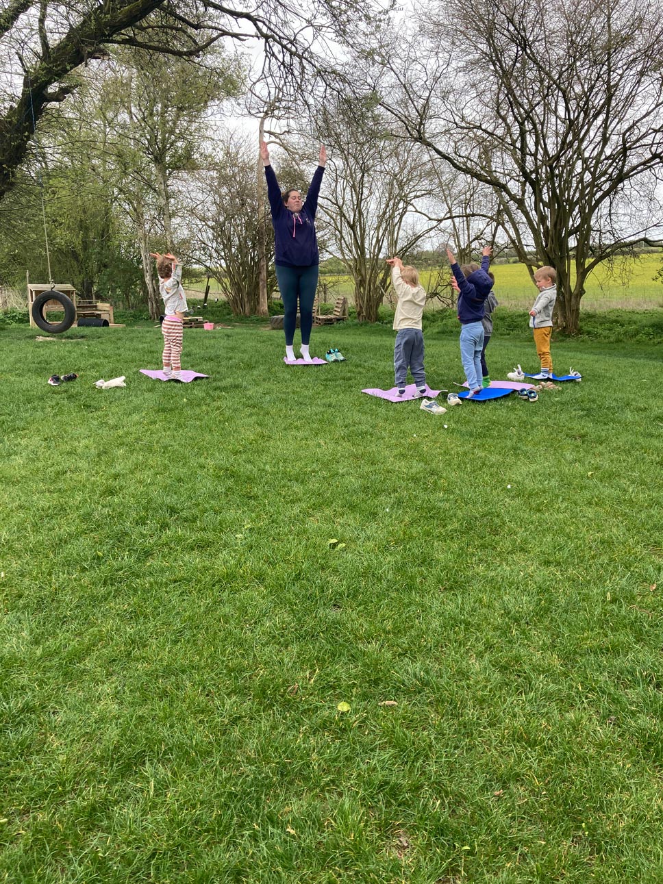 Nursery children following an adult instructor doing arms-up yoga poses outdoors on exercise mats.