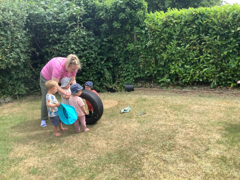 Nursery children in sun hats gathering around a tire with an adult practitioner in a garden area.