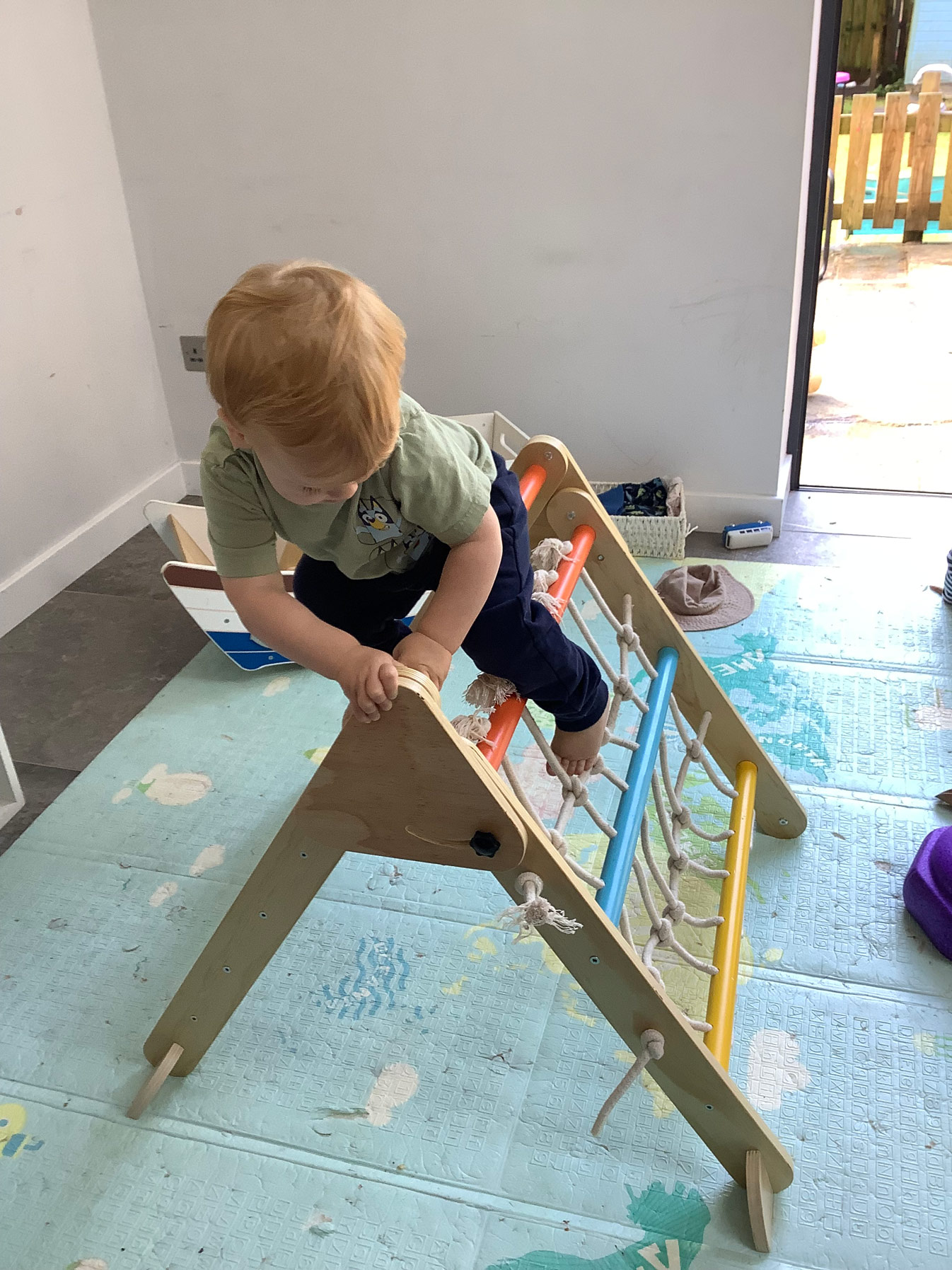 Young nursery child practicing climbing skills and risk-taking on a wooden indoor climbing triangle with a rope net inside the Family Nest