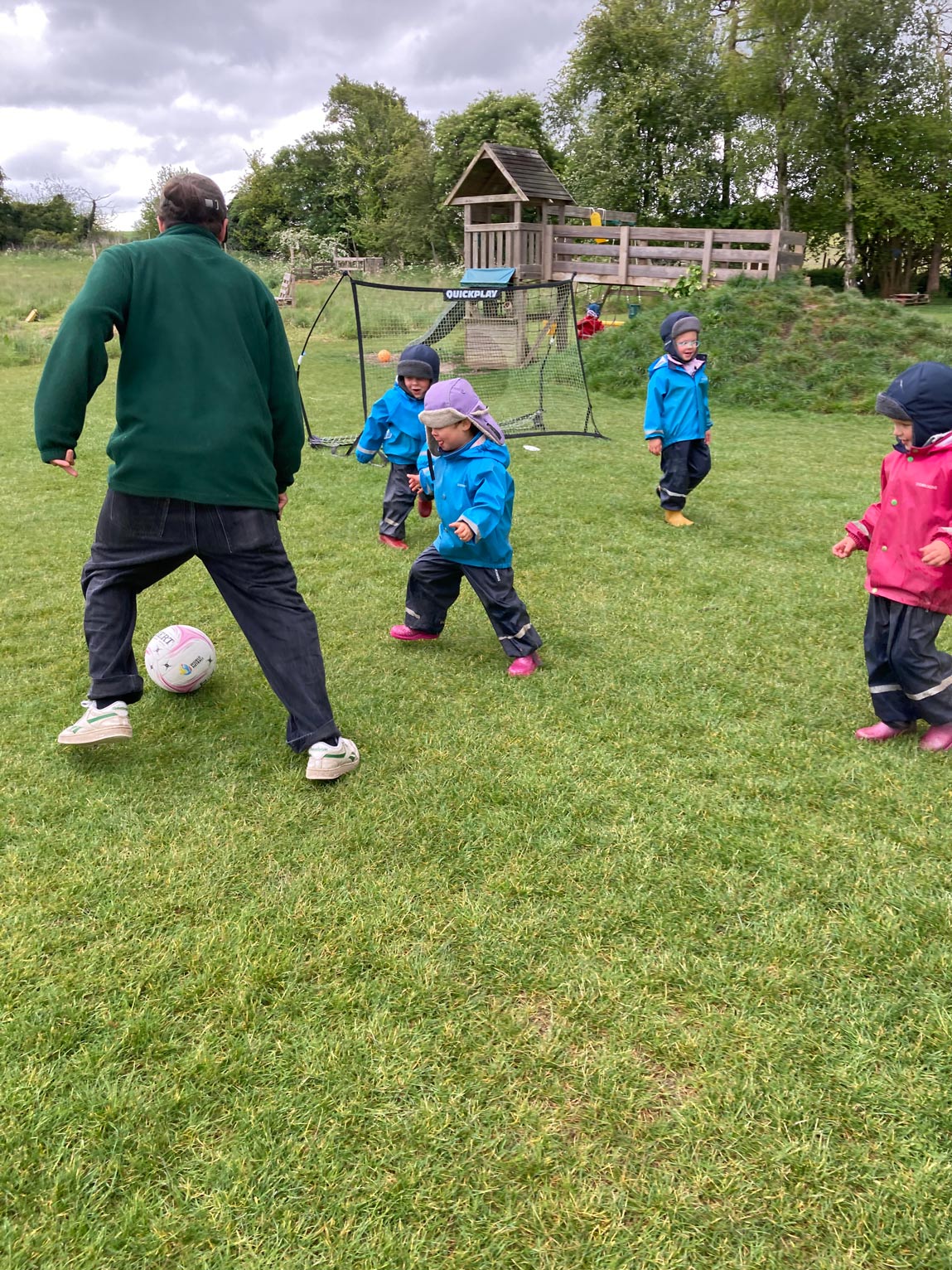 Nursery children in waterproof gear running after a ball during a practitioner-led football game outdoors.