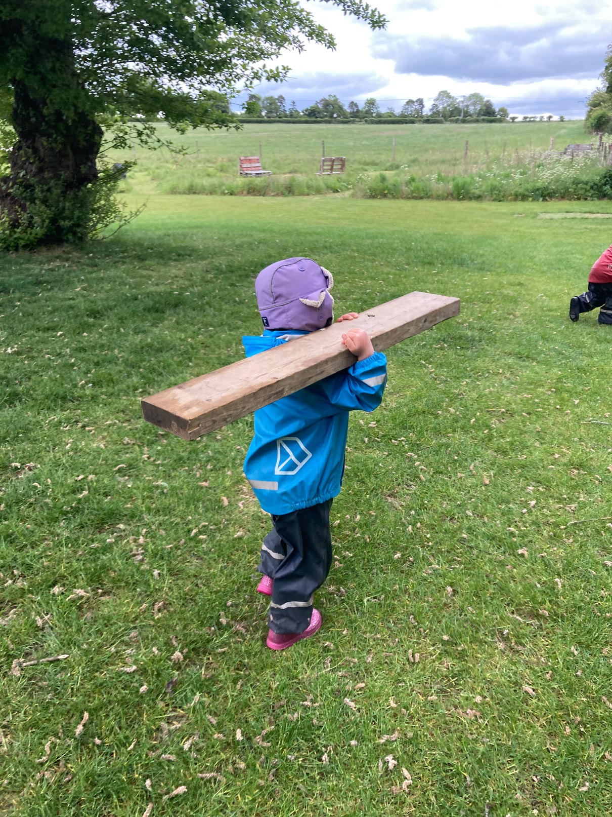 Nursery child in waterproof gear carrying timber for loose parts construction play on a lawn.
