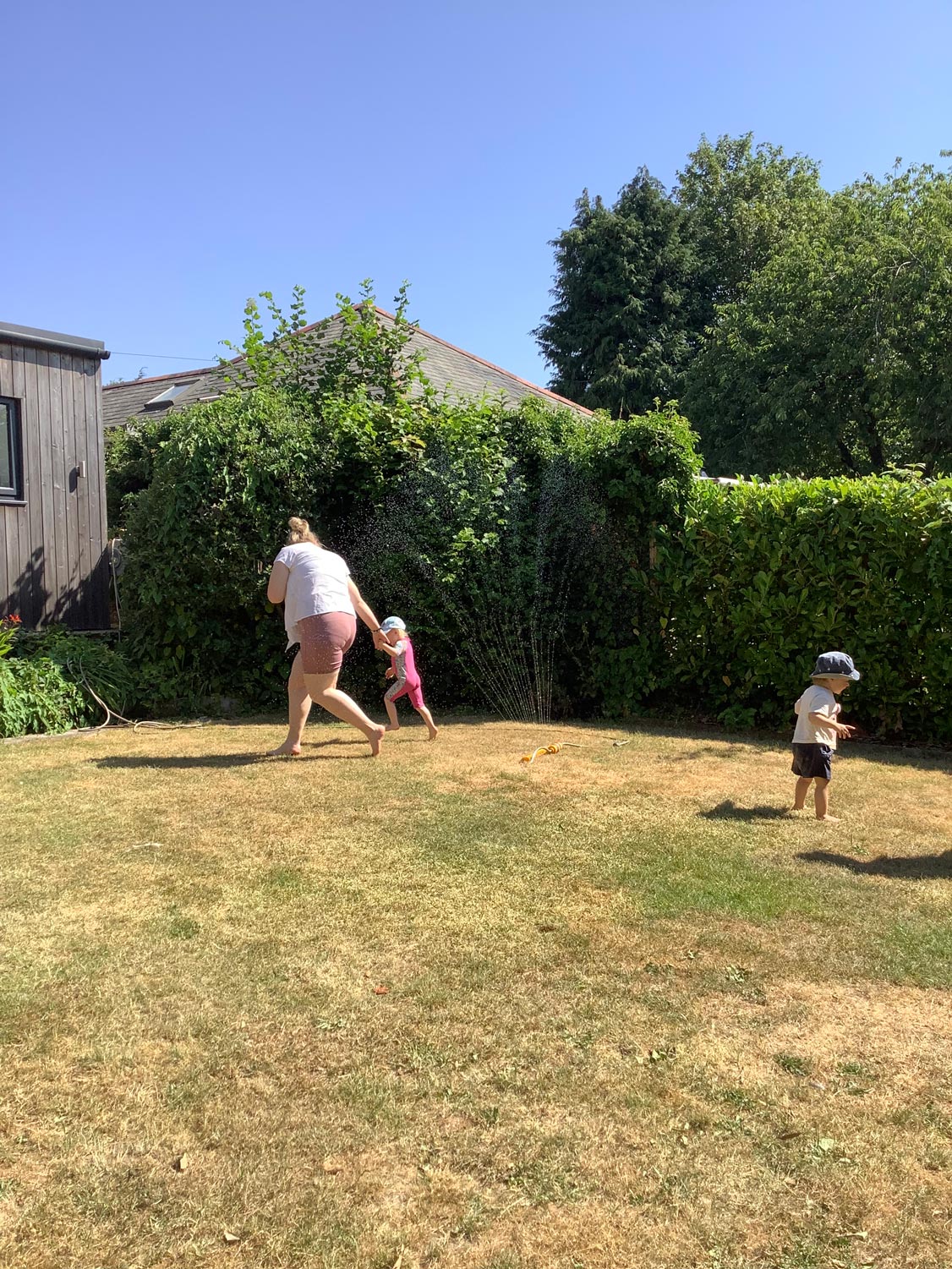 Nursery children playing in a water sprinkler spray outdoors on the lawn under adult supervision.