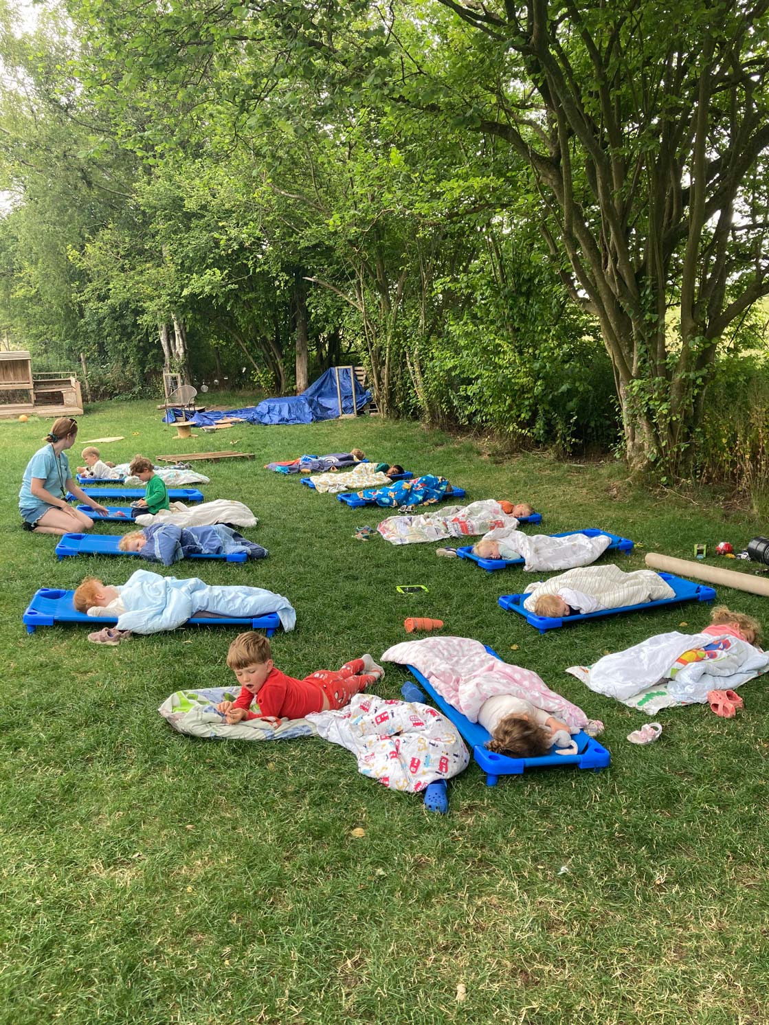 Early years toddlers sleeping on blue nap cots outdoors under tree shade with a nursery practitioner supervising.