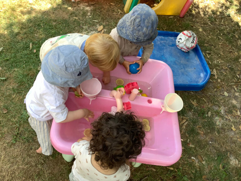 Nursery children exploring floating objects, cups, and citrus slices in a pink sensory water tray outdoors.
