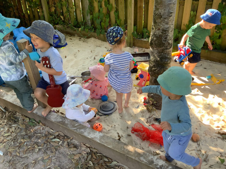 Nursery children playing collaboratively in a shaded outdoor sand pit with toy trucks, scoops, and musical instruments.
