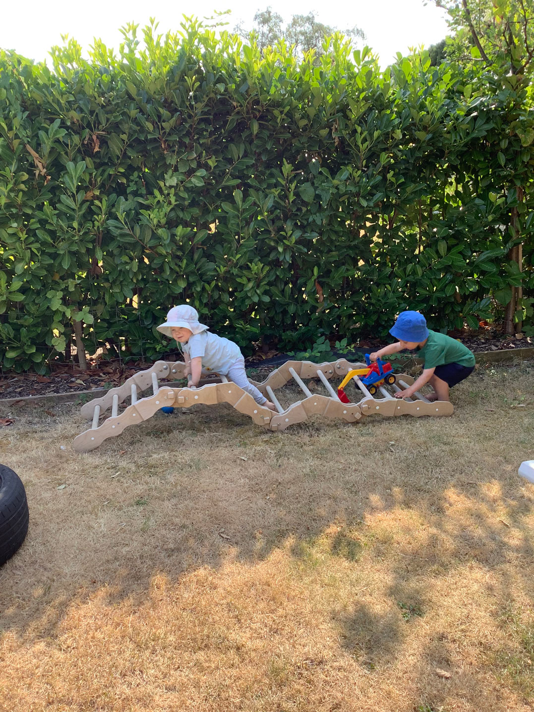 Early years toddlers in sun hats climbing a wooden arch bridge frame and pushing a toy truck outdoors.