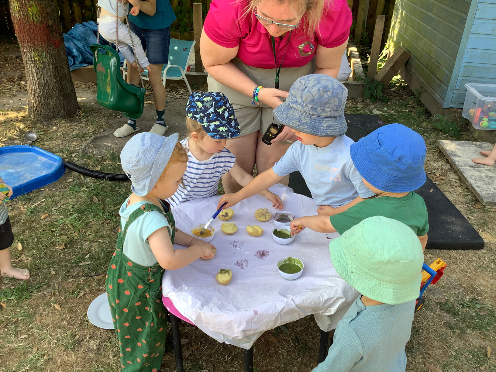 Early years toddlers wearing sun hats using paintbrushes and sliced potatoes for outdoor vegetable printing craft.