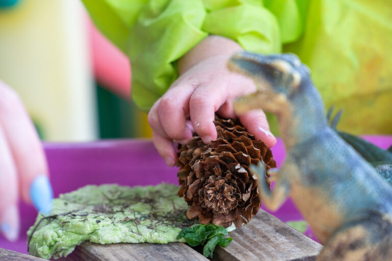 Close up of a child's hand gripping a pinecone with a dinosaur toy in the foreground.