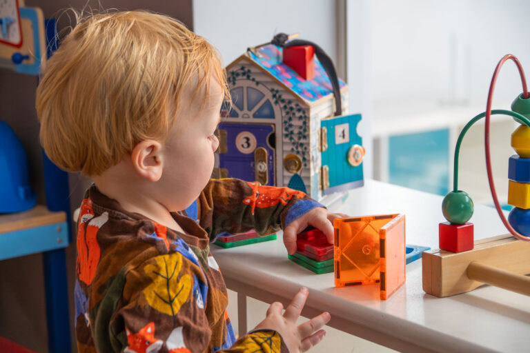 Focused toddler playing with magnetic building tiles alongside a wooden latch house and bead maze toy.