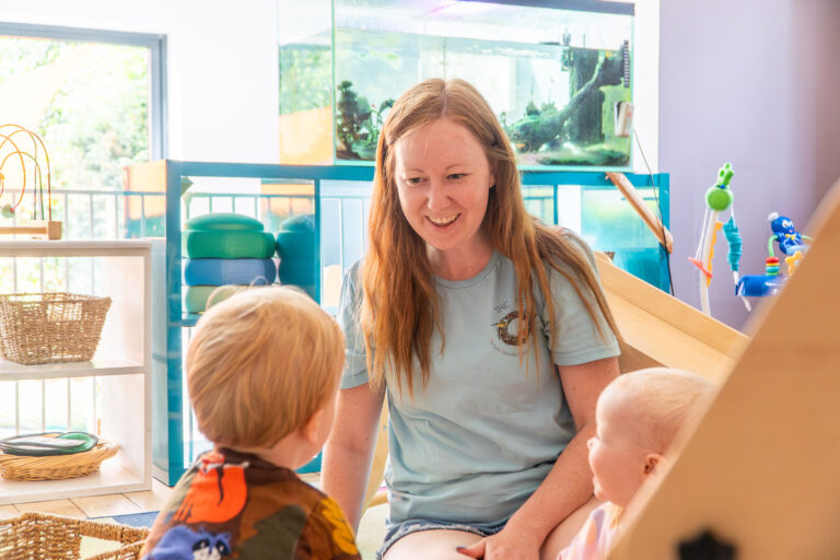 Early years practitioner smiling and talking with two young toddlers in a bright nursery room with a fish tank in the background.