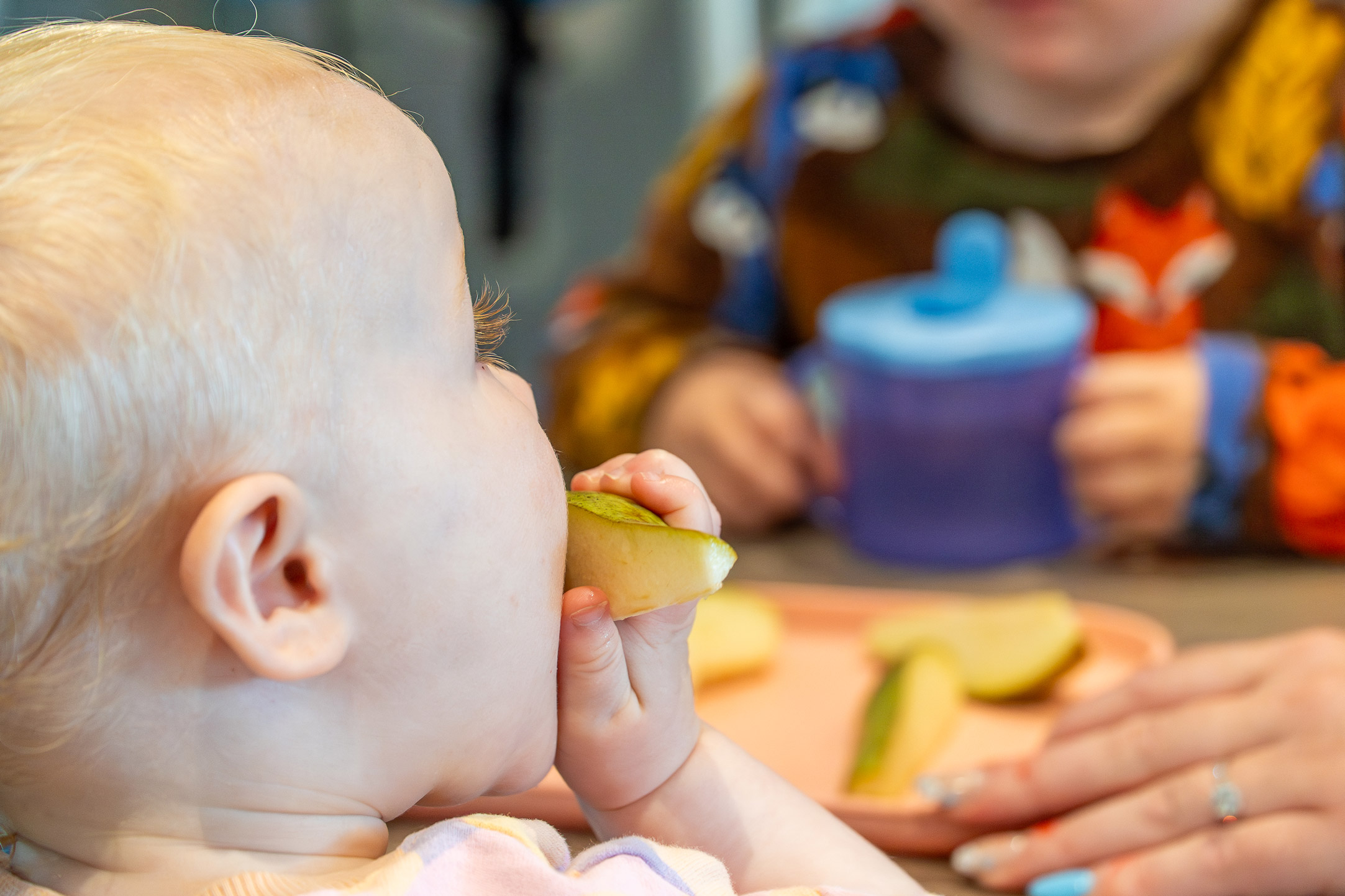 Baby eating fresh fruit slices at a table alongside another child with a sippy cup in the background.